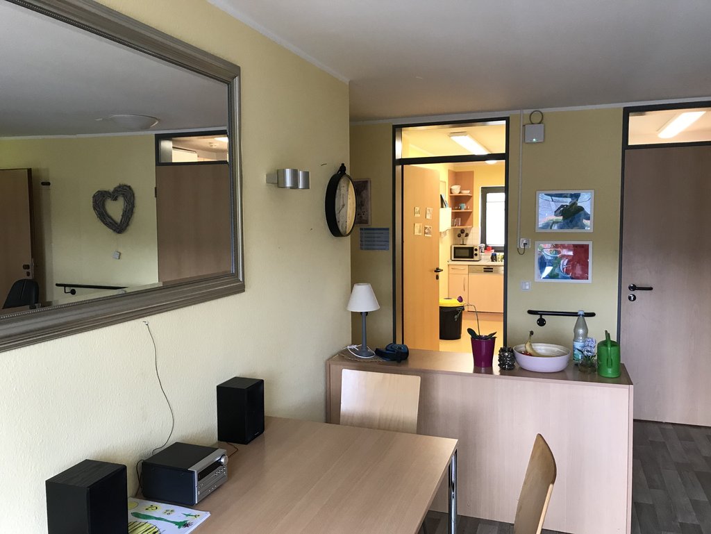 Ein Raum mit einem Tisch, zwei Stühlen, einer Musikanlage, einem Spiegel an der Wand, einer Uhr, einer Lampe und Blick in eine Küche; auf dem Sideboard stehen eine Schale mit Obst, eine Flasche und weitere Gegenstände.