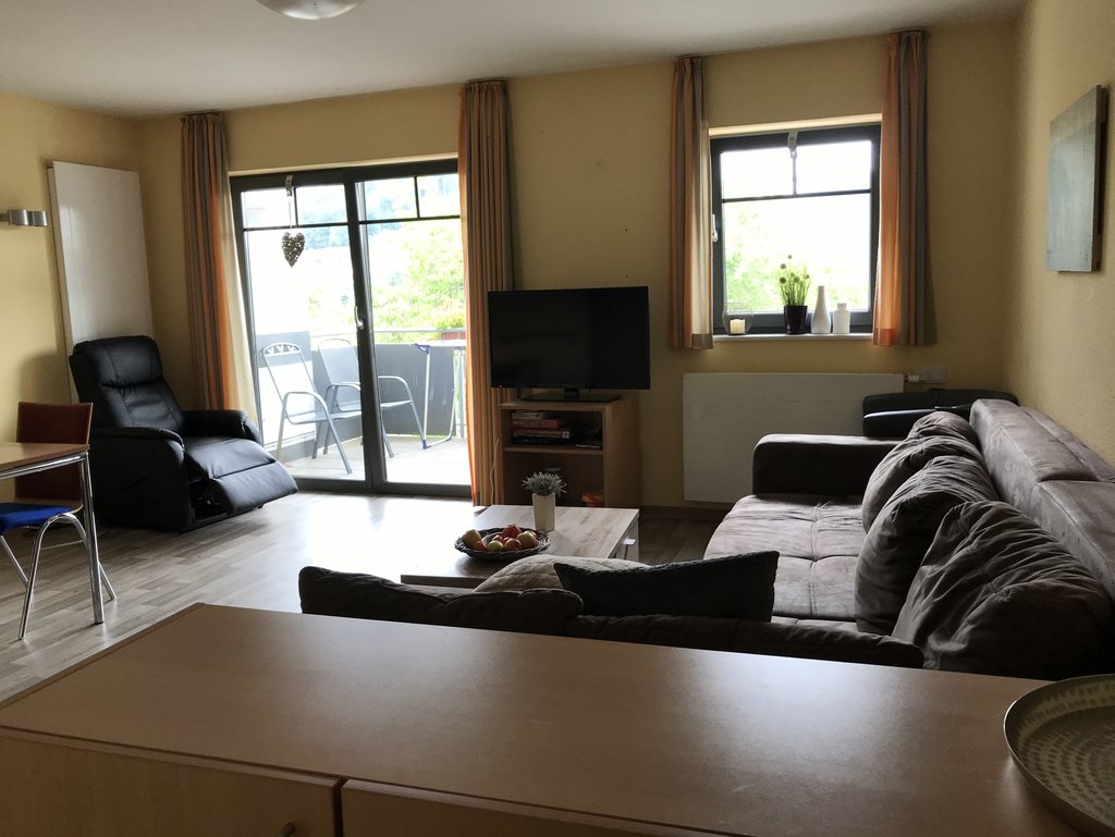 Wohnzimmer mit Ecksofa, Sessel, Couchtisch mit Obstschale, Fernseher auf einem Regal, Esstisch mit Stühlen und Zugang zu einem Balkon mit zwei Stühlen.